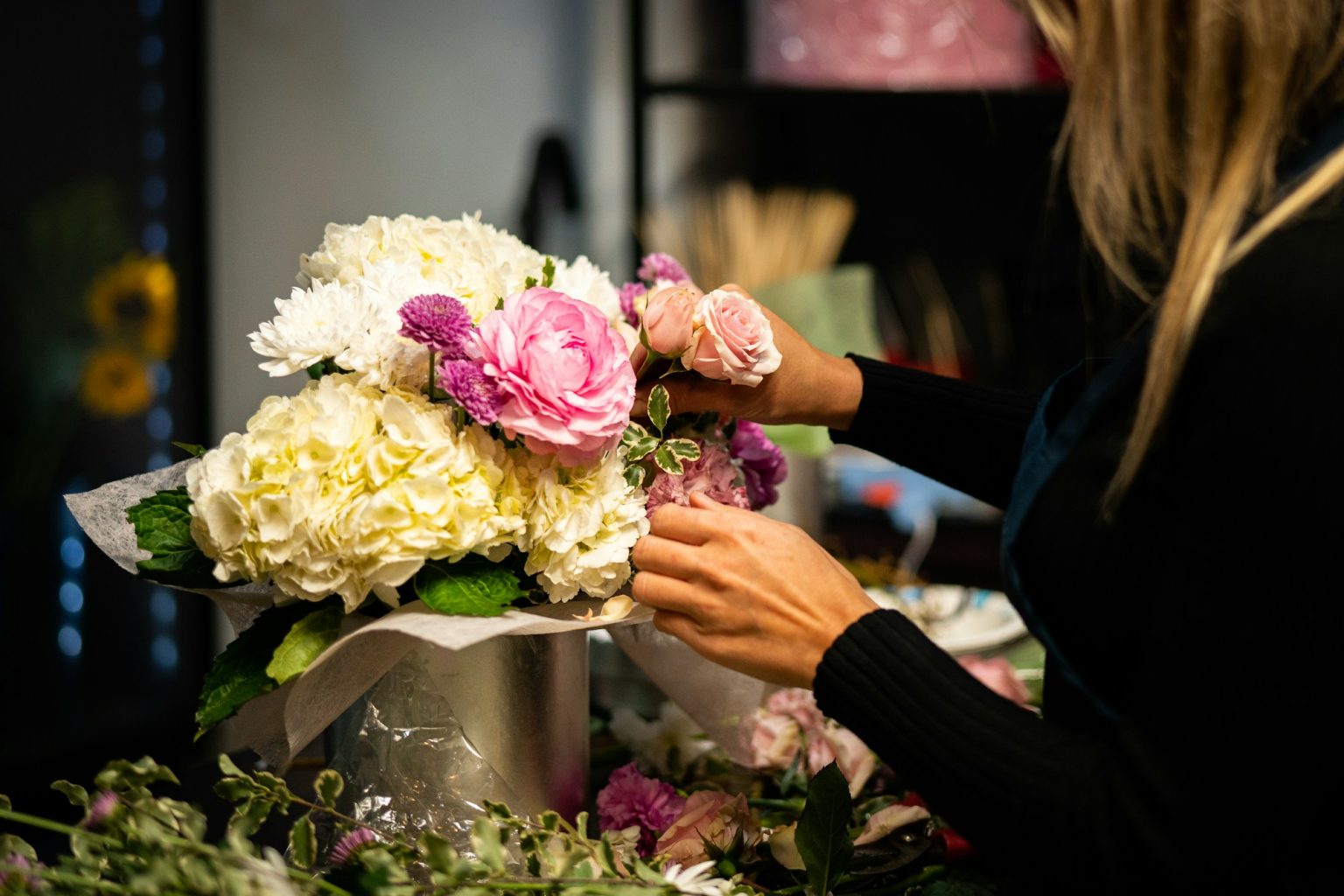 A woman's hands actively arrange stems into a vase of flowers on a table.