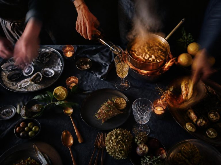 Overhead view of a dark dinner table in motion with wine being poured, a copper pan steaming, oysters on ice, candlelight, and blurred hands.