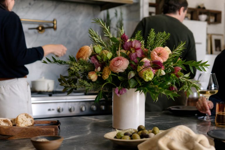 Loose kitchen-island bouquet of ranunculus, hellebores, and herbs in a white cylinder, with bread, olives, and people cooking behind