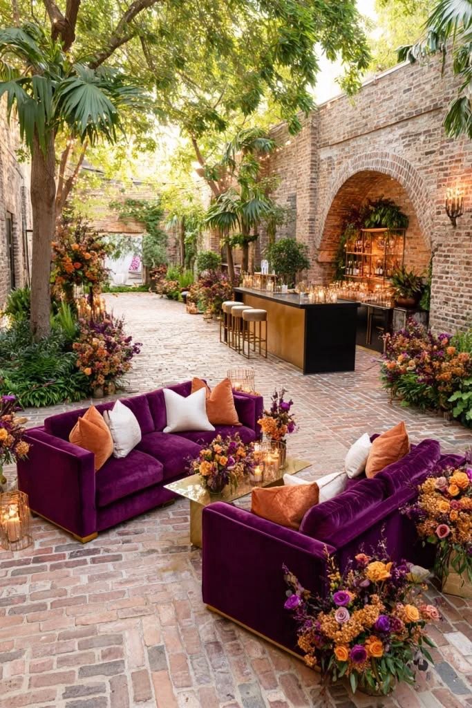 Plum velvet sofas, gold table, and autumn florals in a brick courtyard with a bar under an arch