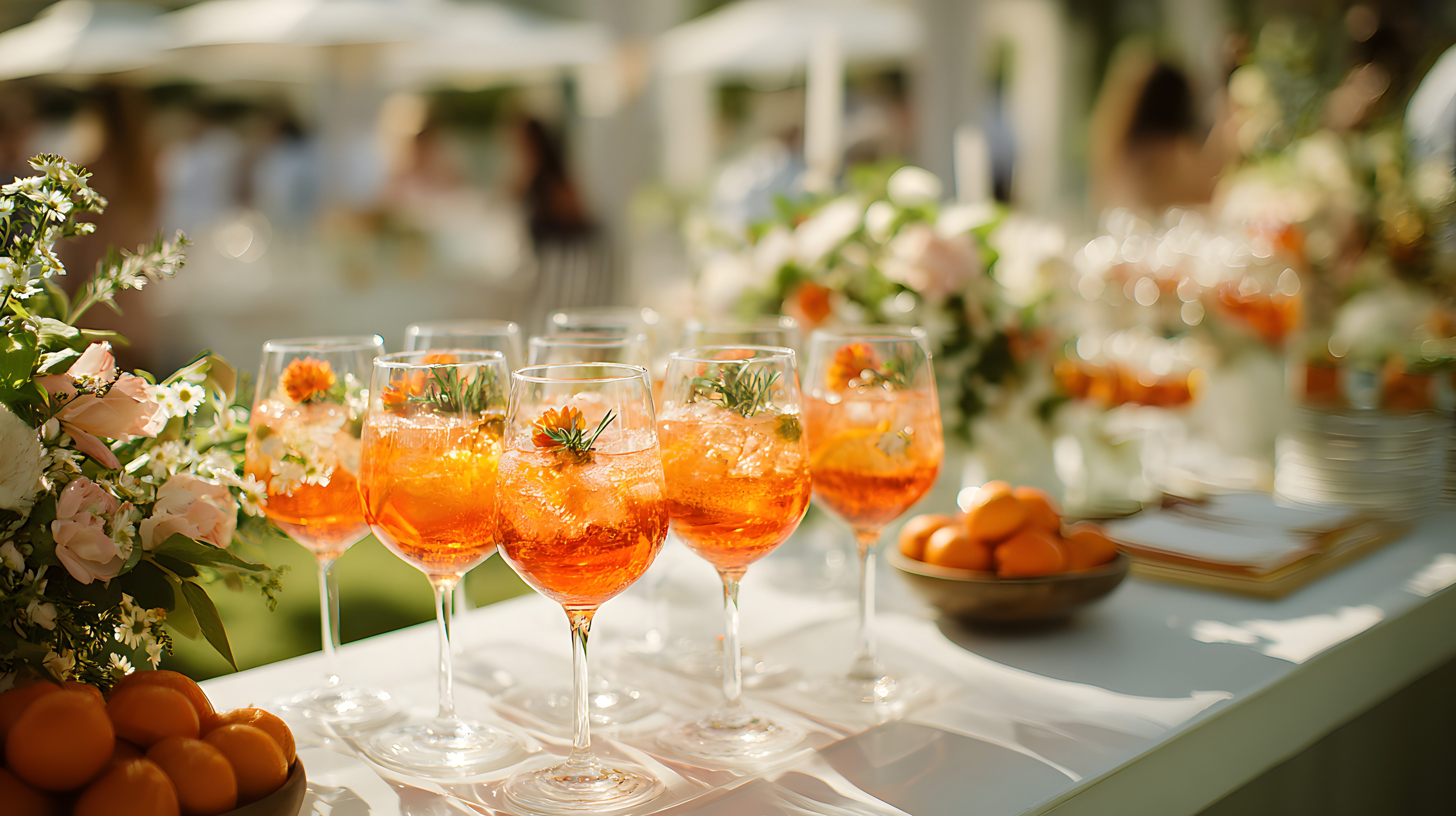 A row of Aperol spritzes poured and waiting on a flower-trimmed welcome table at an outdoor garden reception.