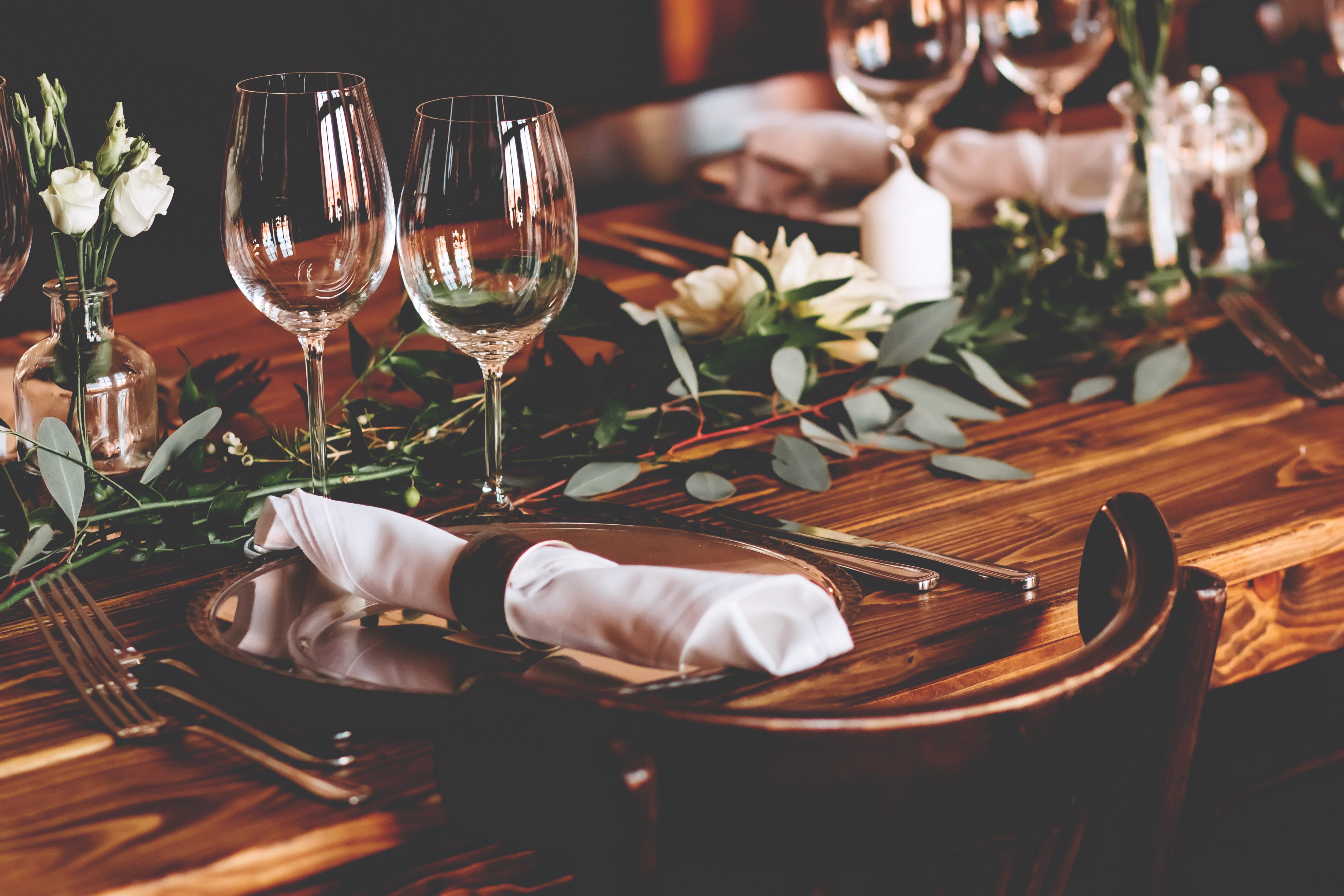 A wooden dinner table with a low run of trailing greenery, small bud vases of white roses and open space between them, set with glassware and folded napkins.