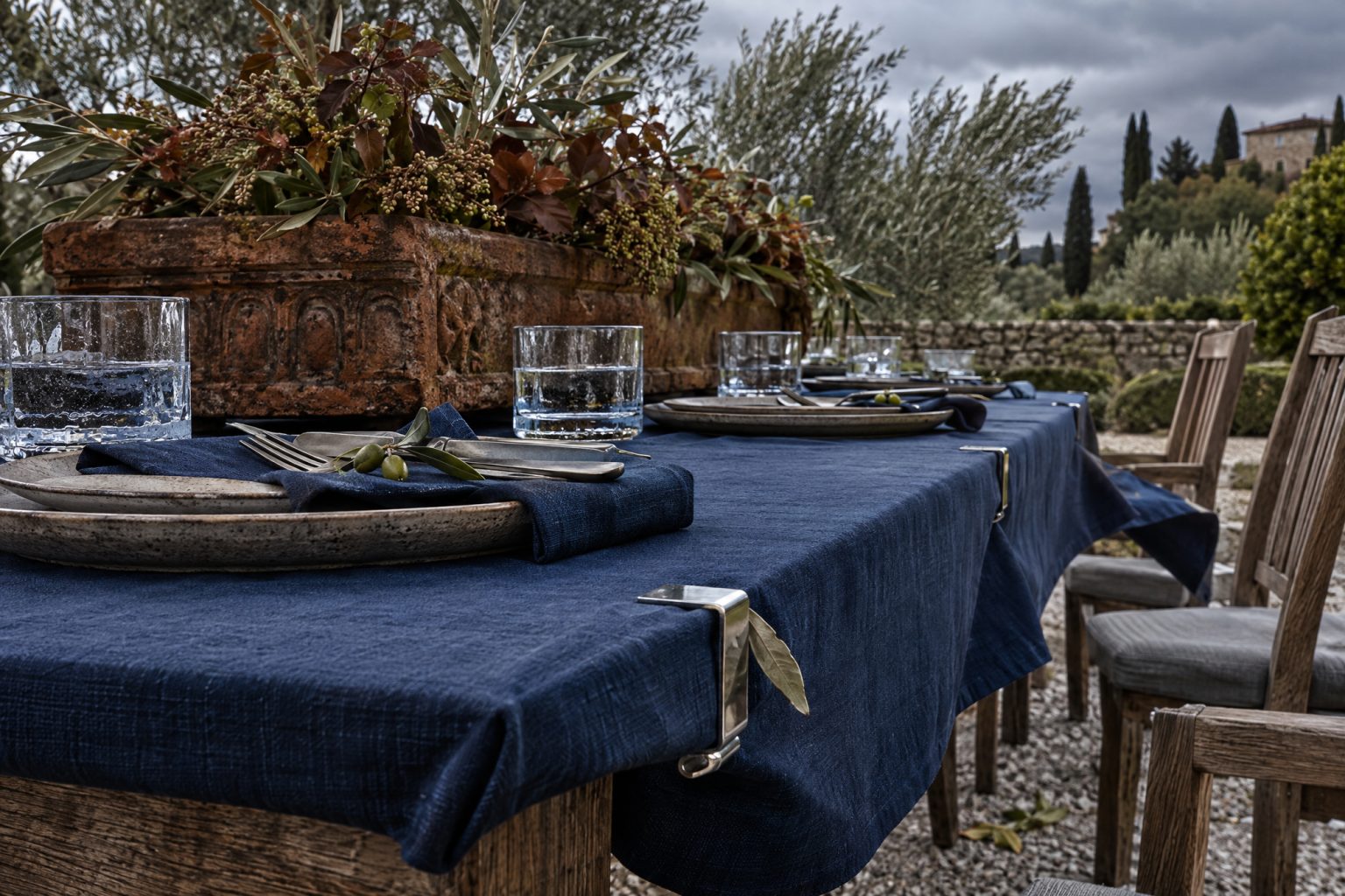 An outdoor table built to survive real weather: steel clips gripping the indigo linen drum-taut to the edge, a carved stone trough too heavy to shift holding low olive and russet foliage, squat thick-based tumblers and weighty stoneware, under heavy overcast rather than golden light.