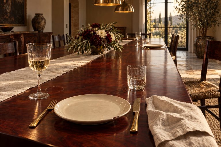 A bare slab of figured mahogany left uncovered and mostly empty: one narrow linen runner laid off-center, hand-blown crystal, heavy gold flatware, a plain plate and a single low arrangement a third of the way down, with long stretches of glowing empty timber between.