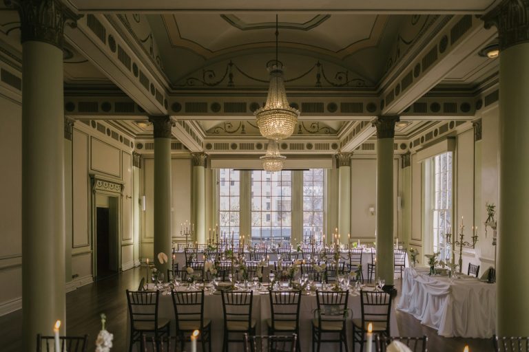 A symmetrical banquet room holds long linen-covered tables and chairs beneath a chandelier.