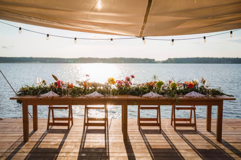 A long dockside dining table with low open floral arrangements and slender glassware keeping the water horizon fully visible.