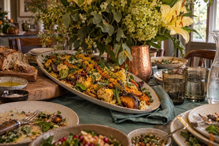 The moment the food arrives and there is nowhere to put it: a heaped serving platter set down tilted half off the table edge because a tall arrangement has taken the middle, its lilies hanging directly over the dish, with plates shoved out of position and glasses touching.