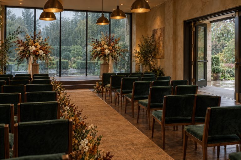 The wet-weather ceremony room set with the same care as the first choice: every chair squarely oriented to the front, arrangements turned to face the seated guests rather than the wall, and the wide accessible doorway left completely clear, with rain running down the windows behind.