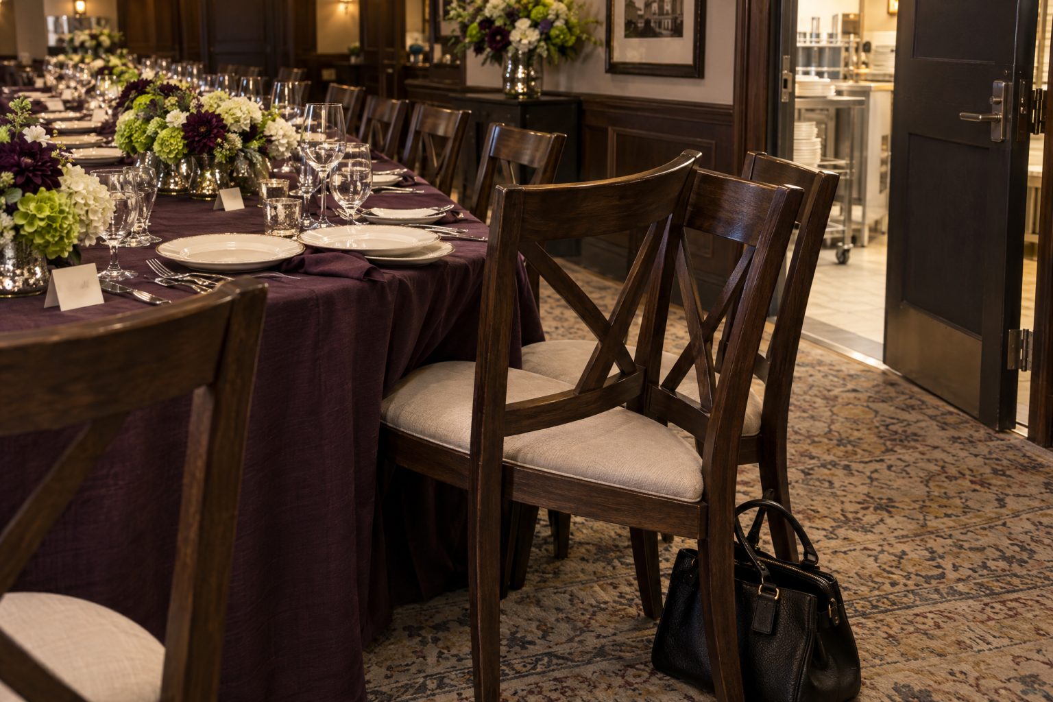 Seen low along a dinner table where the chairs are evenly spaced with room around each setting, until one extra chair is wedged in at an angle in the foreground, overlapping its neighbor with its place setting shoved against the next and a handbag on the floor beside it.