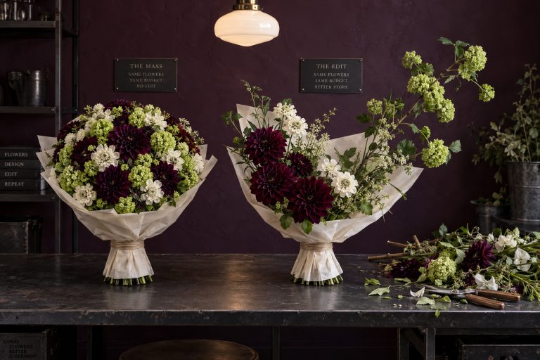 Two bouquets from the same flowers and the same budget standing side by side under identical light: one a tight even dome with no gaps and nowhere for the eye to rest, the other edited to a clear profile with the weight low on one side and a long branch reaching out, its rejected stems discarded on the bench.