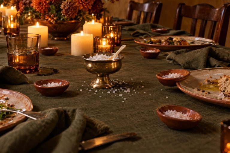 A brass salt cellar with grains scattered round its base sits among pillar candles and amber votives, and at every place along the olive linen a small terracotta pinch dish has already been used — the same small act performed by everyone, with no names anywhere.