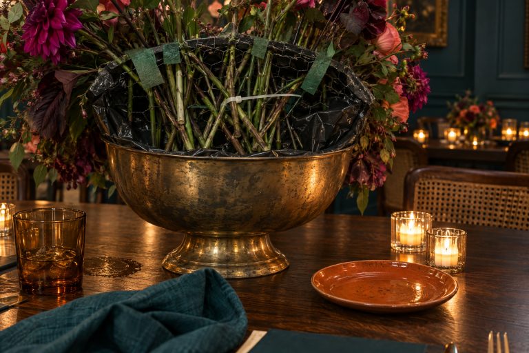 From the seat placed behind it, the unfinished back of a large arrangement in an aged brass bowl — black liner above the rim, chicken wire taped across, bare stems crossing — lit by votives beside the guest's own plate, with every bloom facing away.