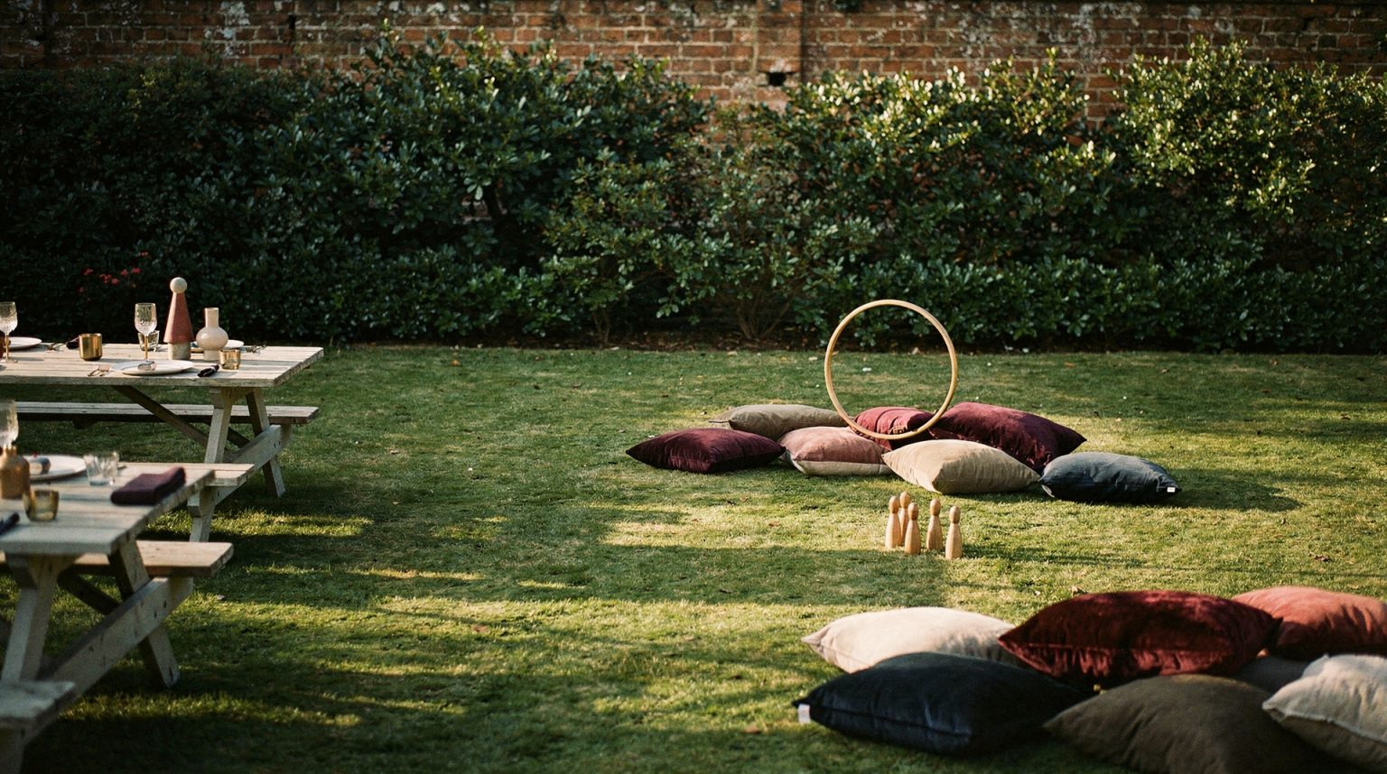 A walled garden lawn in low evening sun with a wooden picnic table at one edge, velvet floor cushions scattered across the grass, and a wooden hoop and a set of skittles left standing in the open middle of the lawn.