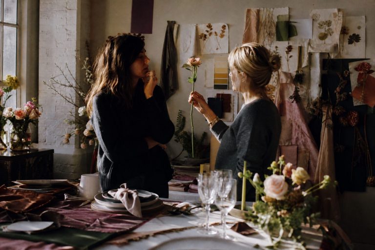 A designer and florist study a stem against a wall of pinned swatches and pressed flowers
