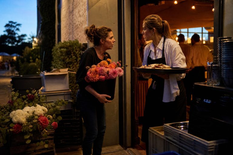 A florist with a bucket of dahlias talks with a catering captain at a venue service door at dusk