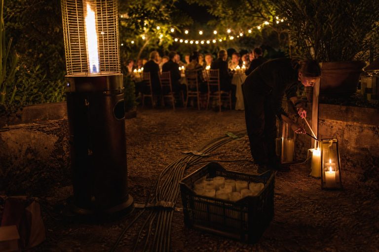 A patio heater, taped cable runs and spare candles in front of a glowing garden dinner under string lights