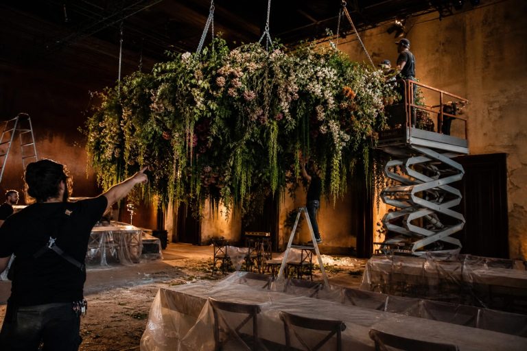 Crew on a lift and ladders raise a huge hanging installation of flowers over a dining room