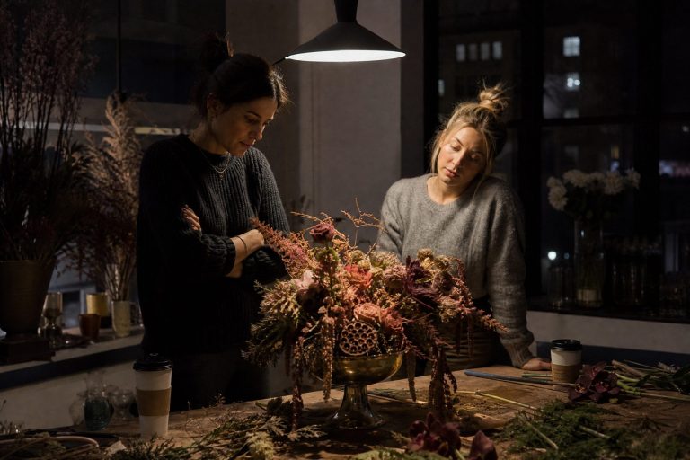 Two women study a sample centerpiece under a pendant lamp in a design studio late at night