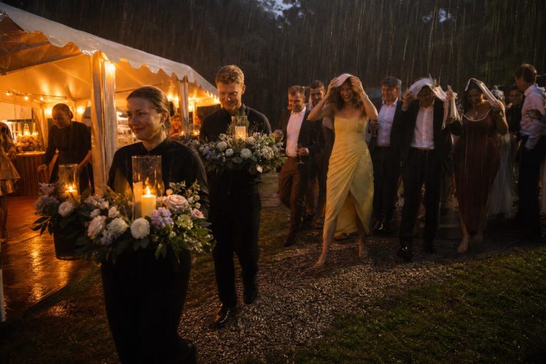 Staff carry flowers and candles into a lit tent as laughing guests follow with their glasses