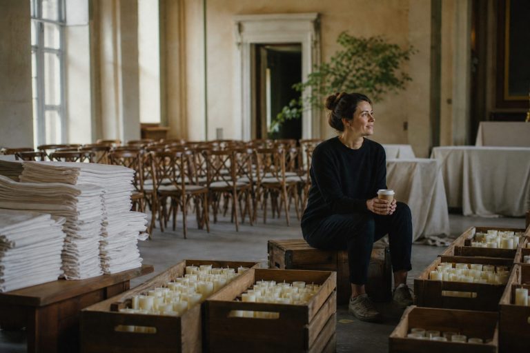 A calm crew member drinks coffee among stacked linens and neat crates of candles in a quiet venue