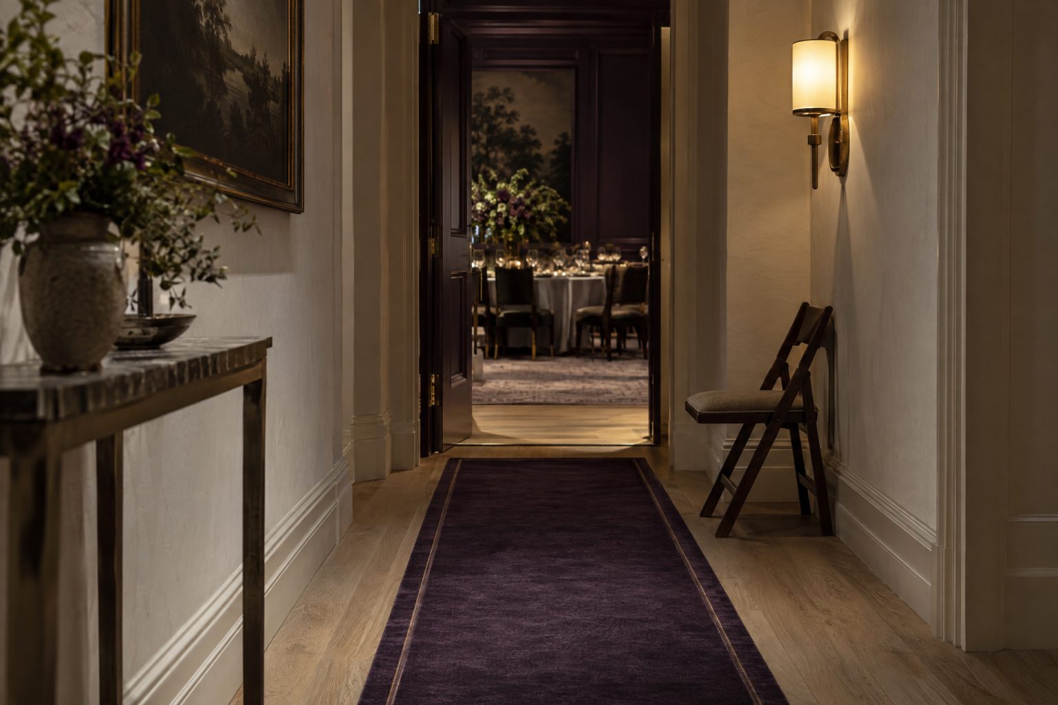 A quiet side corridor with a runner leading to a narrow doorway at the back of a dining room, a low sconce lighting the threshold and a folded chair waiting against the wall for a late arrival to slip in unannounced.