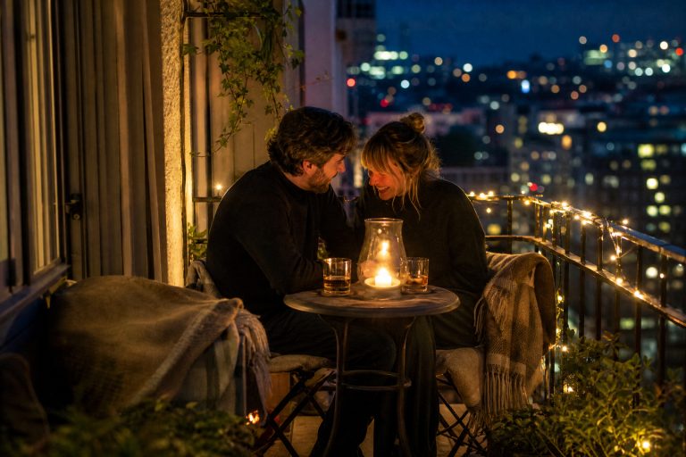 Two people share a nightcap at a small bistro table on a city balcony, wrapped in wool throws with a candle hurricane and fairy lights on the railing.