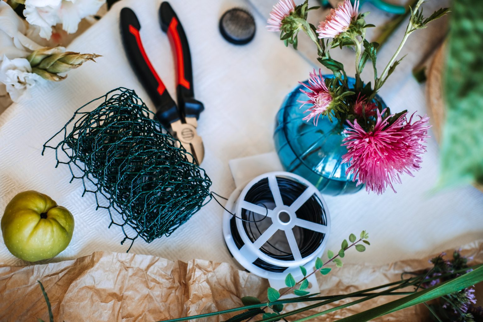 Florist mechanics laid out on kraft paper: a roll of green chicken wire, pliers, a reel of binding wire and tape beside a vase of stems - the structure that holds a loose arrangement together.