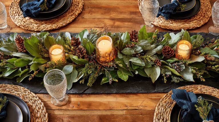 Overhead view of a table run of magnolia leaves, greenery and pine cones with low pillar candles in glass - scent-quiet foliage rather than heavily perfumed blooms.