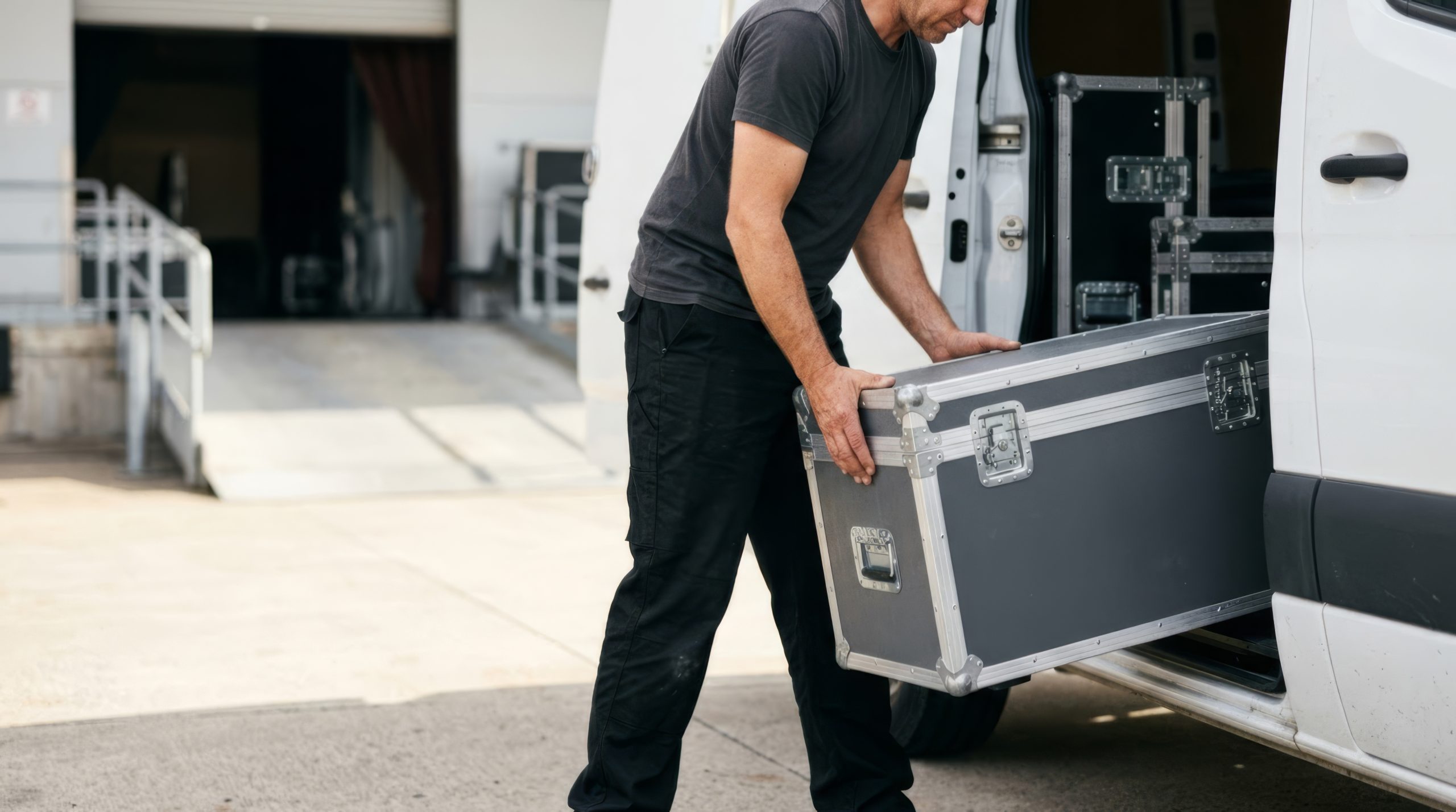 A crew member slides a road case into a cargo van at a venue loading dock, with the dock ramp behind him.