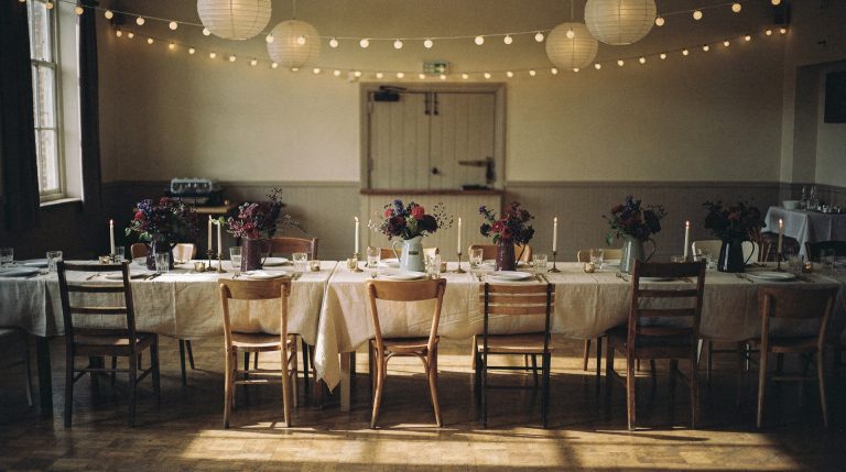 A plain hired hall with a worn parquet floor, laid with one long trestle table under a bone linen cloth, mismatched wooden chairs, jugs of dark flowers and unlit candles down the center, and paper lanterns and festoon bulbs strung overhead.