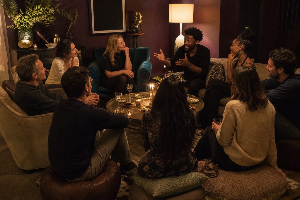 Friends sit close in a circle of sofas, armchairs and floor cushions around a candlelit coffee table in a plum living room, one telling a story with both hands.