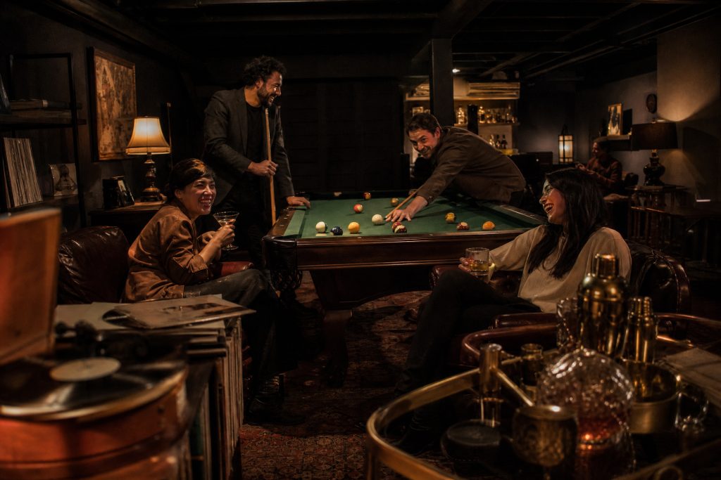 Friends shoot pool and sip cocktails in a low-lit basement den with black beams, leather club chairs, a record player and a brass bar cart.