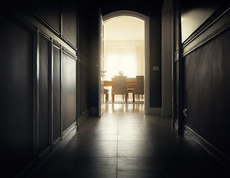 A dark paneled corridor narrowing toward an open doorway, with a bright dining room waiting at the end - the squeeze before the room opens up.