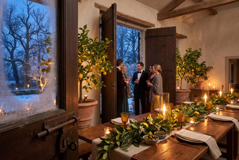 A tall timber shutter swings open mid-dinner to reveal frost-black winter trees beyond the limonaia's glass wall, warm lamplight spilling onto the frost.
